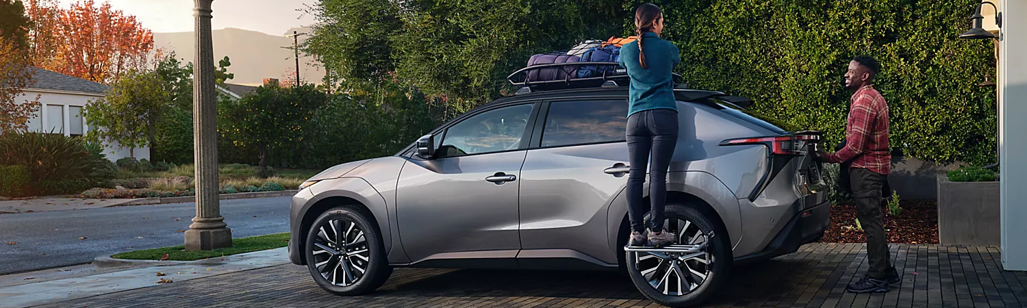 A 2026 Subaru Solterra electric SUV parked in the driveway of a suburban home as a woman stands on a tire step to load the storage attachment on the roof-rails while talking to a man standing at the rear cargo hatch.