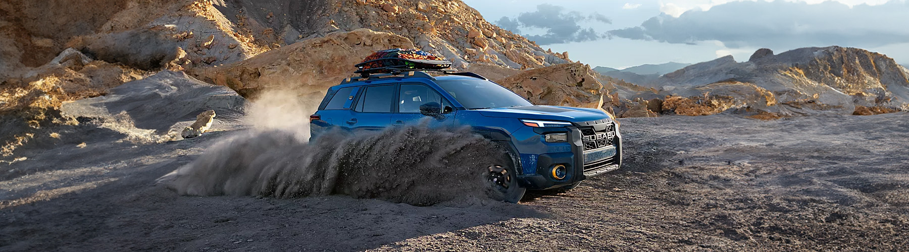 A blue 2026 Subaru Outback with a cargo rack drives on a dirt road in the mountains.