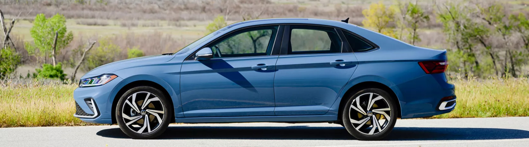 Profile view of a Jetta shown in Monterey Blue Pearl driving with wooded hills in the background