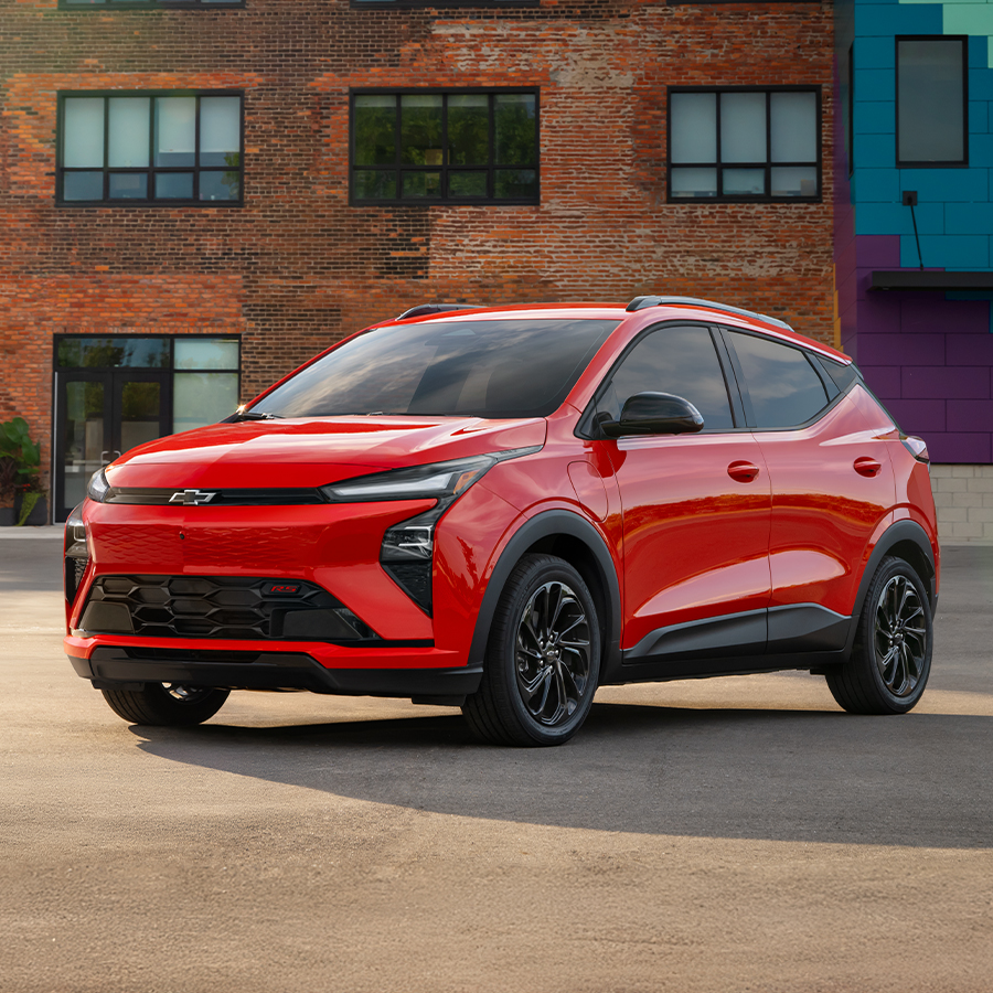 The Chevrolet 2027 Bolt RS exterior is featured in Habanaro Orange (GAG) in a daytime urban setting, from a 3/4 driver's side front angle.