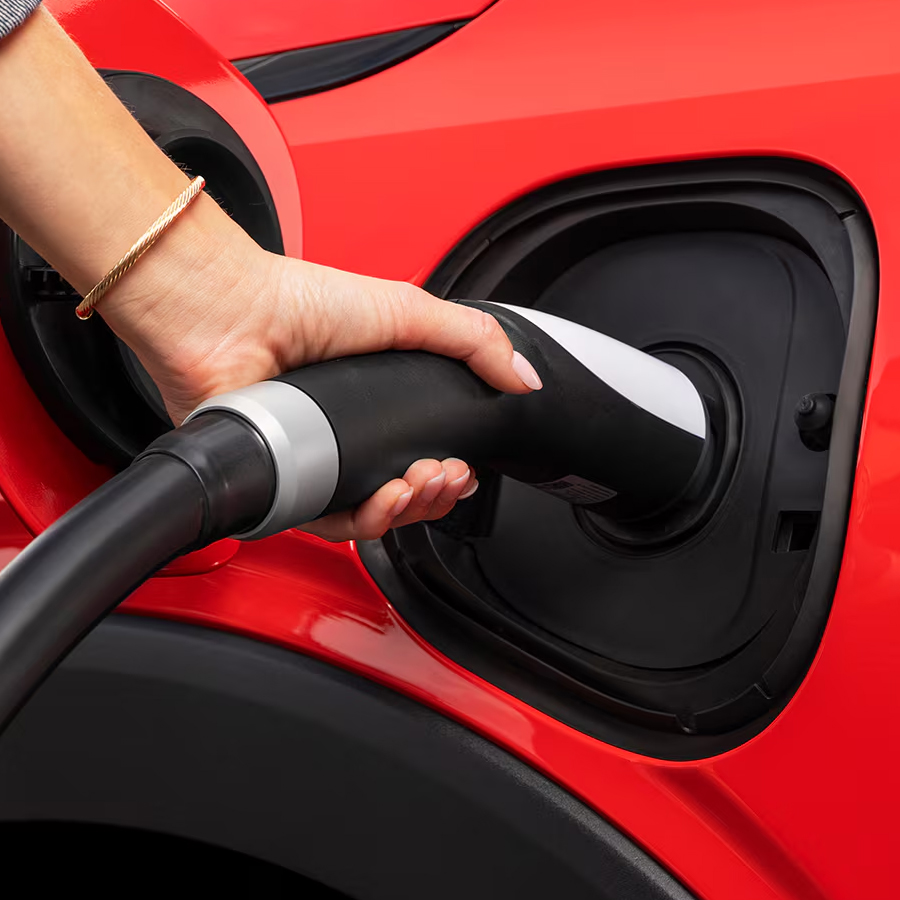 A Close-Up of a Hand Plugging a Charging Cable into the Charging Port of a Red 2027 Chevrolet Bolt.