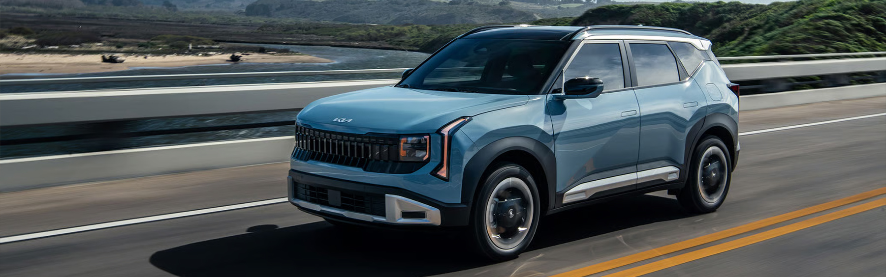 2027 Kia Seltos EX in blue driving on an open highway, shown in front three-quarter view with sleek exterior styling, bold front grille, and a scenic mountain landscape in the background.