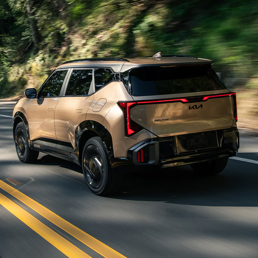 Rear side photograph of the 2027 Kia Seltos in brown as it drives quickly through a hillside forest road, with motion blur distorting the trees and rocky slope behind it. This photo emphasizes the excitement and freedom that driving a Seltos may inspire.
