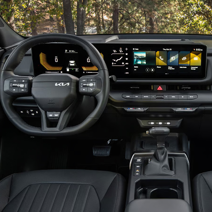 Interior wide-angle photo inside the 2027 Kia Seltos showcasing its steering wheel, dashboard display, center console, shifter, rear view mirrors, glove compartment, and more. A forest is visible through the windshield.