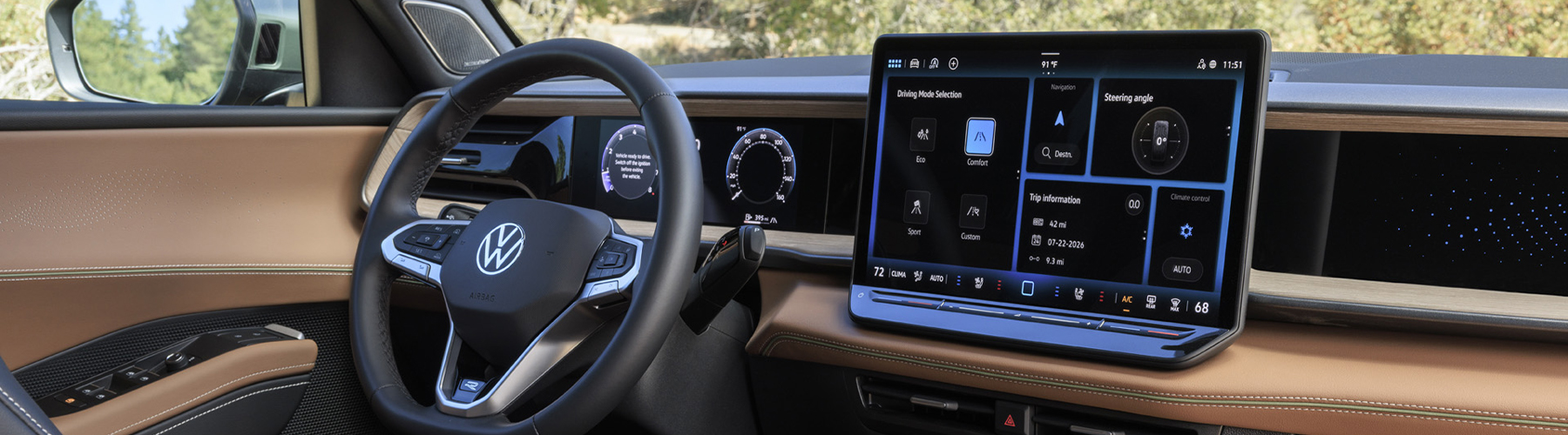 Interior view of an Atlas Cross Sport showing the steering wheel, VW Digital Cockpit and available 15 inch center touchscreen display while trees are visible through the window and windshield.