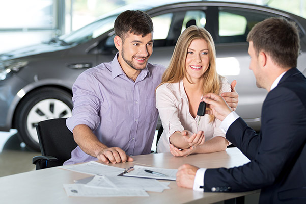 Customers sitting at dealership table with employee handing over keys.