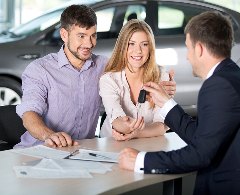 Couple receiving car keys from a salesperson at a dealership desk with paperwork.