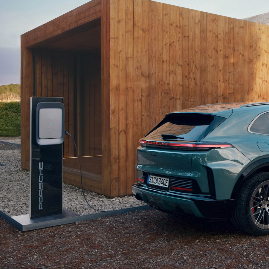 A Porsche Cayenne Turbo is charging at a Porsche Wallbox in front of a building made of concrete and wood.