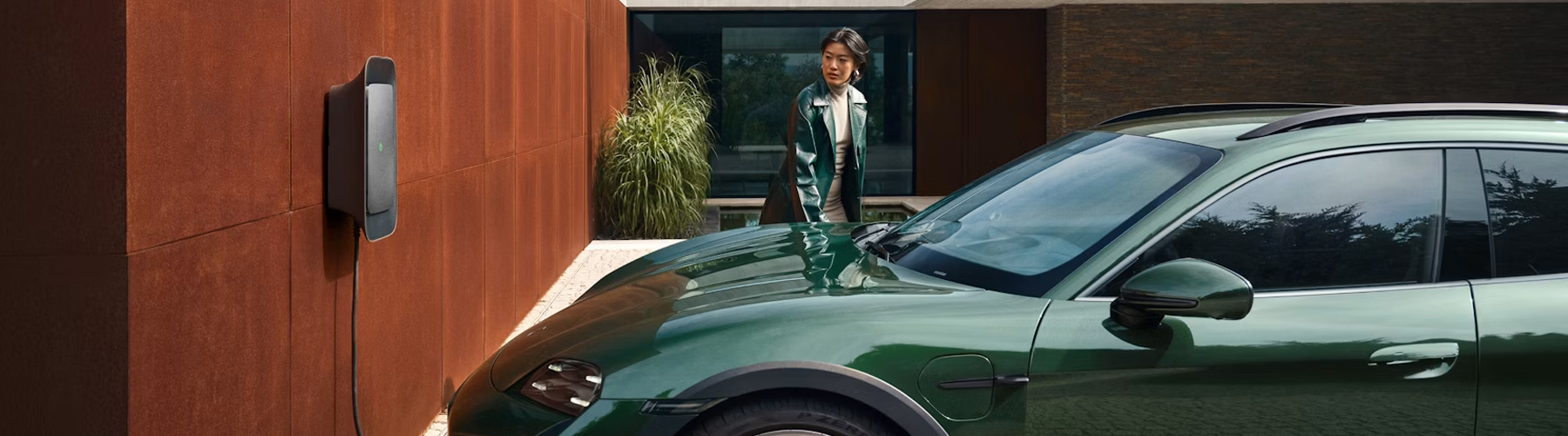 A Porsche Taycan Turbo Cross Turismo in front of a villa next to a Wallbox. A woman plugging in the plug.