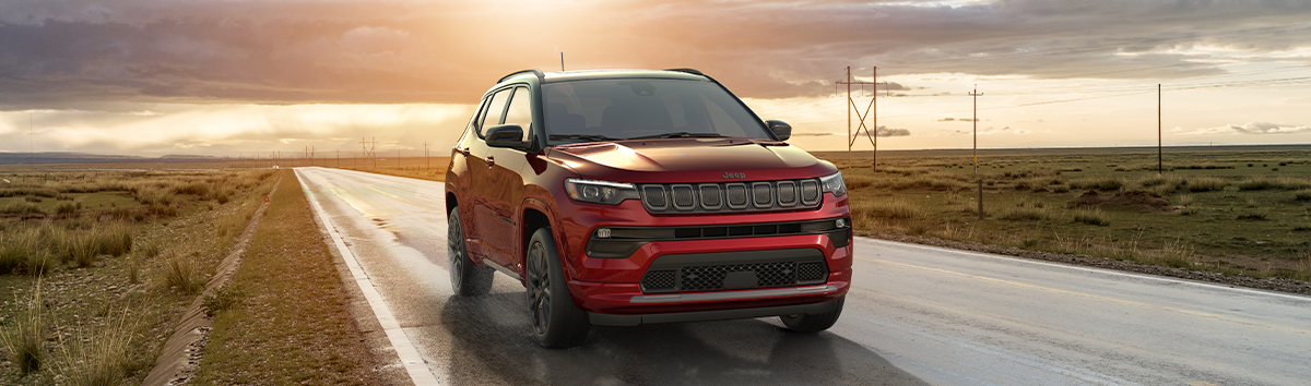 2022 Compass High Altitude in Velvet Red Pearl driving on country highway lined with grass fields, cloudy skies and sun visible in the distance.