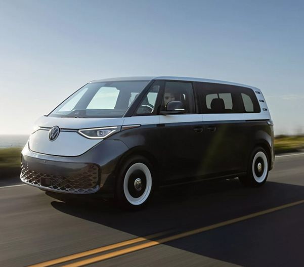 An ID. Buzz shown in Blue Charcoal Metallic and Candy White driving on a brush lined road with the ocean in the background.